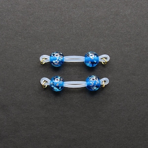 May include: Two pairs of eyeglass retainers. Each retainer features two blue glass beads with white and silver accents, connected by a clear elastic band. Gold-colored metal loops secure the bands.