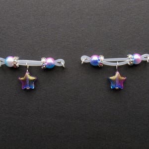 May include: Two nipple rings with a clear band, each featuring a star-shaped charm. The rings have iridescent beads in shades of blue and pink, with silver-colored accents. The star charms are multi-colored.