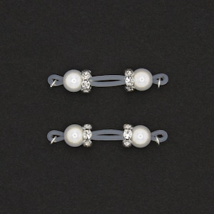 May include: Two pairs of white pearl and rhinestone eyeglass holders on a dark background. Each holder has a white pearl on each end, connected by a clear elastic band and a rhinestone-studded center.