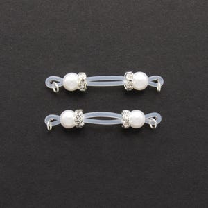 May include: Two pairs of eyeglass retainers. Each retainer features a white pearl bead and a silver-colored, rhinestone-studded accent. The retainers are connected by a clear, flexible cord and have small silver loops at each end.