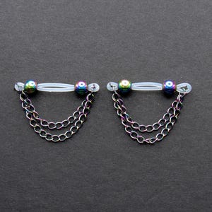 May include: Two nipple rings with iridescent beads and dangling silver chains. The rings have a clear, flexible bar and are set against a dark gray background. The chains are made of multiple strands.