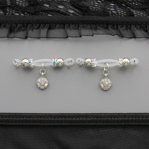 May include: Two nipple rings with clear bands, silver beads, and dangling round, iridescent charms. The background is gray with black lace and fabric at the top and bottom.
