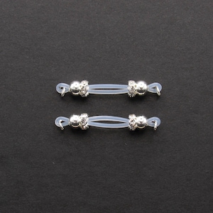 May include: Two pairs of eyeglass retainers. Each retainer features a clear, flexible band with a silver-colored decorative bead at each end. The beads have a floral design and are adorned with small, clear crystals. The background is a solid black.