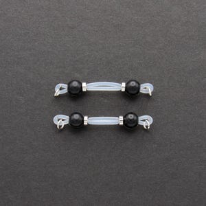 May include: Two pairs of black bead eyeglass retainers. Each retainer features a black bead at each end, connected by a clear, flexible tube. Silver-colored metal loops are attached to the ends for securing to glasses.