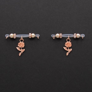 May include: Two nipple rings with a rose gold rose charm. Each ring has a clear band with gold-colored beads and a dangling rose pendant. The background is a solid black.
