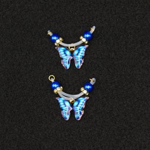 May include: Two butterfly-shaped jewelry pendants. Each pendant features a blue and purple butterfly with a gold-colored frame. The top of each pendant has blue beads and clear plastic accents. The background is black.