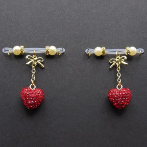 May include: Two nipple rings with a clear band, each featuring a gold bow and a dangling red heart covered in small, sparkling rhinestones. The bands have gold accents and faux pearls. The background is a solid black.