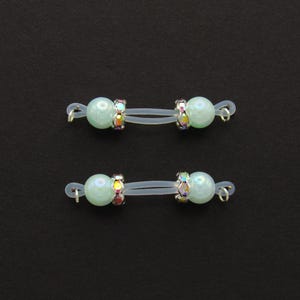 May include: Two pairs of nipple jewelry. Each piece features a pale green pearl bead, accented with iridescent rhinestones and connected by a clear, flexible band. The jewelry has small silver-colored loops on each end.