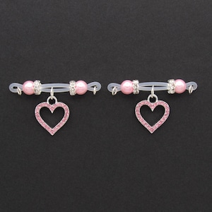 May include: Two nipple rings with a clear band, pink heart pendants, pink beads, and silver accents. The heart pendants are encrusted with pink rhinestones. The background is black.