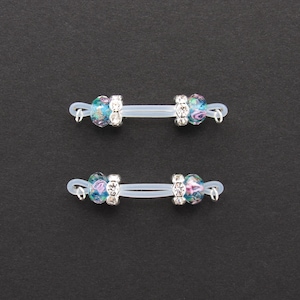 May include: Two nipple rings with clear, flexible bars. Each ring features a central section with clear rhinestones and colorful glass beads in shades of blue, pink, and green. The background is a solid black.