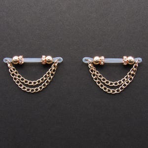 May include: Two gold-colored nipple chains with a clear connecting bar. Each chain features a small, round bead with a cluster of small crystals and two strands of delicate chain. The background is a solid black.