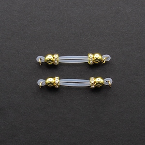 May include: Two pairs of eyeglass retainers. Each retainer features a clear, flexible band with gold-colored decorative accents. The accents include a gold bead with clear rhinestones and a small gold loop for attachment to glasses.
