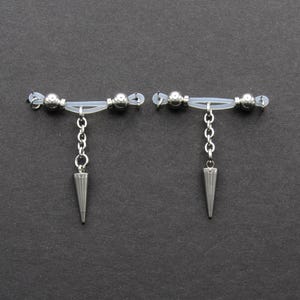 May include: A pair of silver-toned earrings with a unique design. Each earring features a horizontal bar with clear beads and a dangling chain ending in a cone-shaped pendant. The earrings are set against a dark gray background.