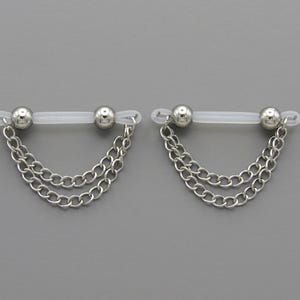 May include: Two silver-colored nipple rings with a chain design. Each ring features a clear, flexible bar with a silver ball on each end. Two strands of chain hang from each bar, creating a draped effect.