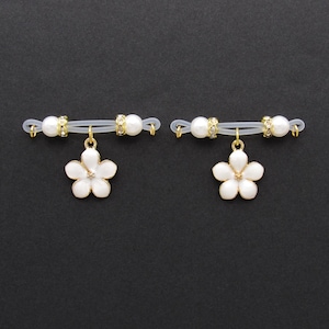 May include: Two white flower nipple pasties with gold-colored accents and a pearl and rhinestone design. The pasties are connected by a clear elastic band. The background is black.