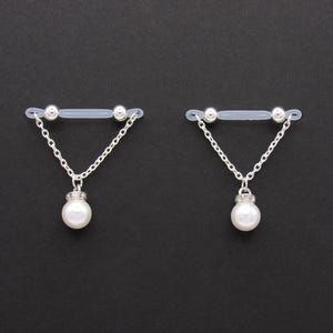 May include: Two nipple jewelry pieces. Each piece features a clear, flexible bar with silver-colored beads at each end. A silver chain hangs from the bar, connecting to a pendant with a white pearl and silver accents. The background is black.