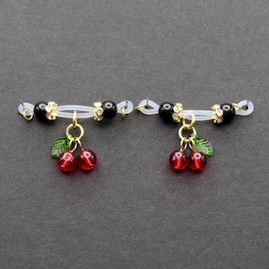 May include: A pair of nipple jewelry featuring a clear band with black beads, gold accents, and dangling red cherry charms with green leaves. The jewelry is designed to be worn on the nipples.