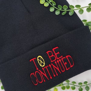 May include: Black knit beanie with red embroidery that reads "TO BE CONTINUED" with a yellow circle and an X inside.