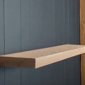 May include: A light wood shelf mounted on a dark gray wall. The shelf is made of unfinished wood and has a natural grain pattern.