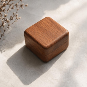 Puede incluir: Una pequeña caja de madera cuadrada de color marrón intenso. La caja tiene una superficie lisa y pulida con esquinas redondeadas. La tapa encaja perfectamente en la base. La caja es probablemente para joyas o pequeños recuerdos.