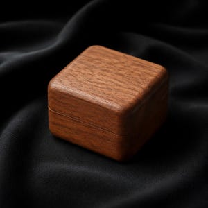 Può includere: Una scatola quadrata di legno con coperchio incernierato. La scatola è realizzata in legno marrone scuro con un motivo a venature visibili. La scatola è appoggiata su una superficie nera e strutturata.