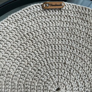 May include: Close-up of a beige crocheted item with a textured surface. A small brown leather tag with the word "Handmade" is attached. The item appears to be a hat or a similar accessory.