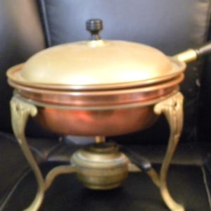 May include: A vintage copper and brass chafing dish with a lid and a black handle. The dish has ornate, gold-colored legs and a burner base. The lid is a light gold color, and the dish itself is copper.