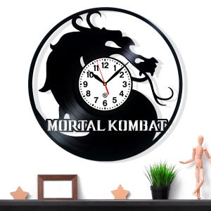 Könnte beinhalten: Schwarze Vinyl-Schallplattenuhr mit einer Drachensilhouette und dem Schriftzug "MORTAL KOMBAT". Das Zifferblatt ist weiß mit schwarzen Zahlen und Zeigern. Eine einzigartige Wanduhr für Videospiel-Fans.