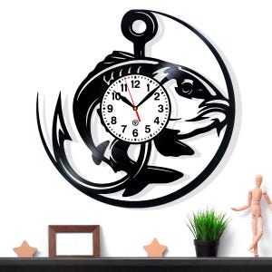 May include: A black wall clock with a fish design, crafted from a vinyl record. The clock face is white with black numerals and hands. The fish is set within a circular frame, with a hook at the top. The clock is mounted on a white wall.