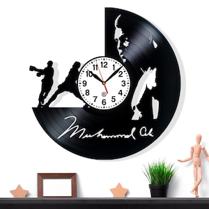 Könnte beinhalten: Schwarze Vinyl-Schallplattenuhr mit einer Boxer-Silhouette und einem Porträt. Das Zifferblatt ist weiß mit schwarzen Zahlen und Zeigern. Die Uhr enthält die Unterschrift "Muhammad Ali".