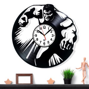 May include: Black vinyl record clock featuring a silhouette of a muscular figure with a clock face at the center. The clock hands are black and red. The clock is mounted on a white wall.