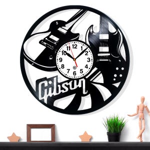 May include: Black and white wall clock with a guitar design. The clock face is round with black numbers and hands. The word "Gibson" is displayed in white script. The clock is made from a vinyl record.
