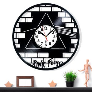 May include: Black vinyl record clock featuring a white clock face with black numbers and hands. The clock has a black triangle design and the words "Pink Floyd" in a stylized font. The clock is surrounded by a brick-like pattern.