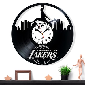 May include: Black vinyl record clock featuring a basketball player silhouette dunking above a cityscape. The clock face is white with black numbers and hands. The Los Angeles Lakers logo is displayed at the bottom.