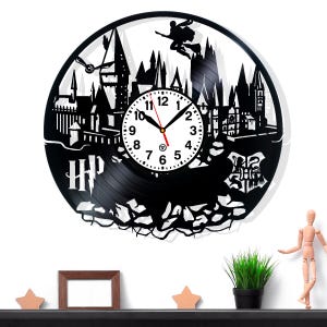 Puede incluir: Reloj de vinilo negro con la silueta de un castillo, una figura voladora y las letras "HP". La esfera del reloj es blanca con números y manecillas negras. Un artículo decorativo para fans.