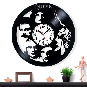 Puede incluir: Un reloj de pared en blanco y negro hecho de un disco de vinilo, con la banda Queen. La esfera del reloj es blanca con números y manecillas negras. La palabra "QUEEN" está en la parte superior. El reloj está montado en una pared blanca.
