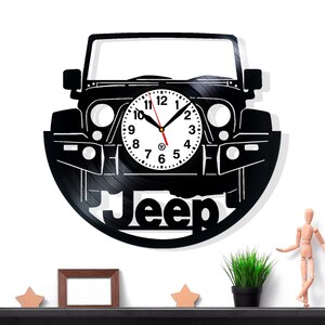 Può includere: Orologio da parete nero a tema Jeep. Il quadrante dell'orologio è bianco con numeri e lancette nere. L'orologio è tagliato da un disco in vinile nero a forma di Jeep. La parola "Jeep" è ritagliata in basso.