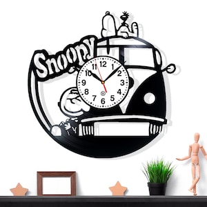 Può includere: Orologio da parete nero a tema Snoopy. L'orologio presenta Snoopy e Woodstock su un design di autobus VW. Il quadrante dell'orologio ha numeri e lancette nere. L'orologio è montato su una parete bianca.
