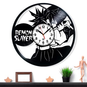 Op de afbeelding: Zwarte en witte Demon Slayer vinylplatenklok. De klok heeft een karakterillustratie met een zwaard en de woorden "Demon Slayer". De wijzers van de klok zijn zwart en rood.
