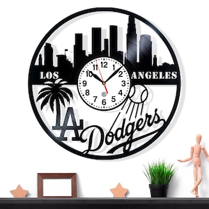 May include: Black Los Angeles Dodgers wall clock. The clock features a cityscape silhouette, a palm tree, and the Dodgers logo. The clock hands are red. The clock is approximately 12 inches in diameter.