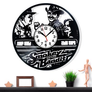 以下が含まれることがあります： 「Smokey and the Bandit」のキャラクターが描かれた黒いビニールレコード時計。時計の文字盤は白で、黒い数字と針が付いています。映画のタイトルは、その下に白い文字で表示されています。