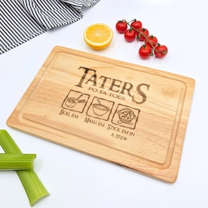 Könnte beinhalten: Ein rechteckiges Schneidebrett aus hellem Holz mit einer Lasergravur, die Taters Po-ta-toes sowie Illustrationen und den Text Boil em, Mash em, Stick em in a stew zeigt. Das Brett verfügt über eine umlaufende Saftrille.