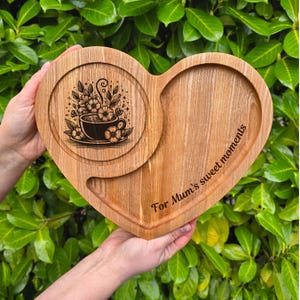 Può includere: Un vassoio di legno a forma di cuore con un inserto circolare con un disegno floreale di tazza da tè. Il vassoio è inciso con le parole "For Mum's sweet moments".