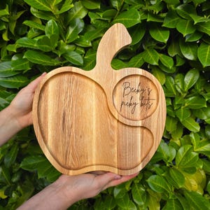 May include: Wooden apple-shaped serving board with a handle. The board has a circular indentation and a smaller, curved indentation. The text "Becky's picky bits" is engraved in the circular indentation. The board is held against a backdrop of green leaves.