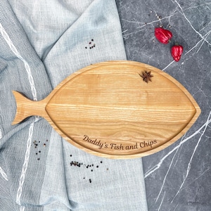 Puede incluir: Una tabla de servir de madera con forma de pez, con el texto "Daddy's Fish and Chips" grabado. La tabla marrón claro está sobre un paño a rayas azules y blancas. Hay pimientos rojos y especias esparcidas.