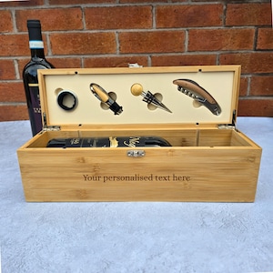May include: A rectangular bamboo wine box with a hinged lid featuring the engraved text "Your personalised text here". The interior holds a wine bottle, a drip ring, a pourer, a bottle stopper, and a corkscrew. A wine bottle stands behind.