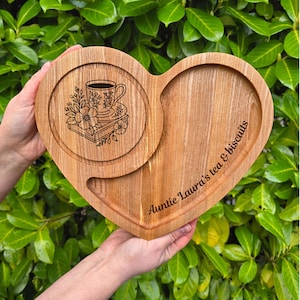 Può includere: Vassoio di legno a forma di cuore con un'area sottobicchiere circolare con un disegno floreale di tazza da tè. Il vassoio ha un'area incassata per biscotti e il testo "Auntie Laura's tea & biscuits".