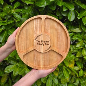 May include: A round, wooden serving tray with three compartments and a central circular area. The tray is light brown with a natural wood grain. The center has the text "The Hughes' Nibbly Bits" within a decorative border.