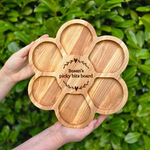 May include: A wooden serving board in a flower shape, featuring six compartments. The board has the text "Susan's picky bits board" in the center. The board is held by hands against a backdrop of green foliage.