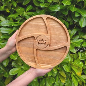May include: A round, wooden serving board with four sections. The board is light brown with a natural wood grain. The center of the board has the text "Emily's board" in a circle. The board is held by two hands against a backdrop of green leaves.